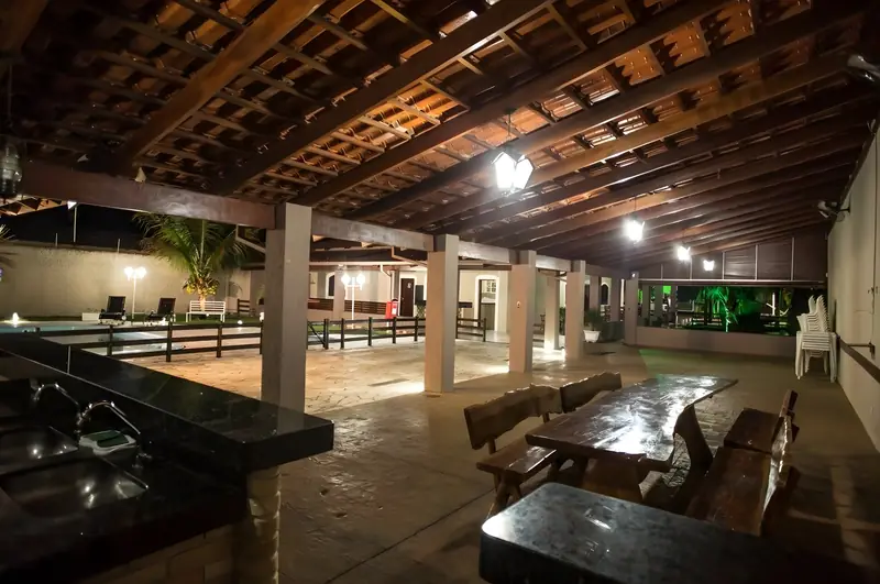Área gourmet à noite, com bancada de granito, mesas de madeira e a piscina iluminada ao fundo