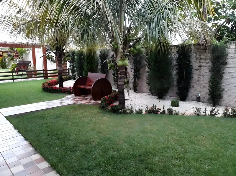 Jardim com gramado, coqueiros, canteiro de flores e banco decorativo vermelho
