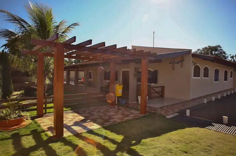 Pérgola de madeira no jardim, com gramado, coqueiro e a casa ao fundo