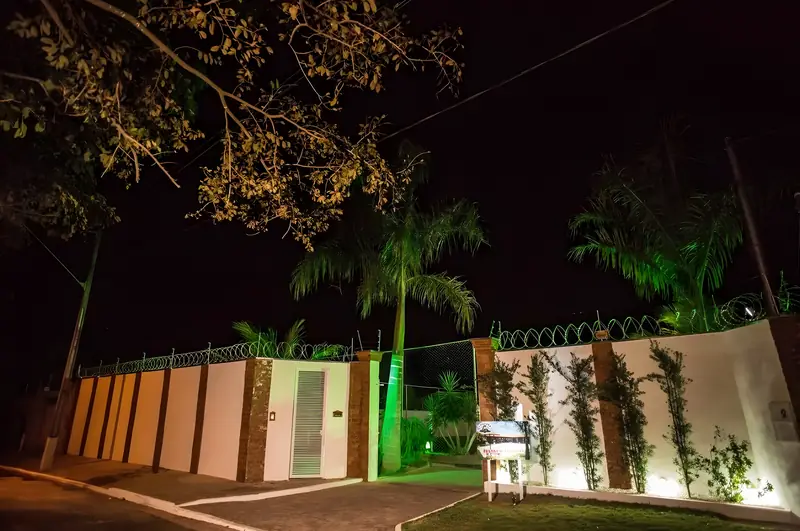 Portão de entrada da Chácara Império iluminado à noite, com palmeiras e muro com trepadeiras